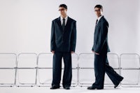 two men in suits standing in front of chairs